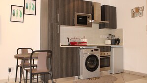 Una cucina compatta con lavatrice, forno a microonde e un piccolo frigorifero. È presente un tavolo da pranzo con sedie e opere d'arte incorniciate appese al muro.