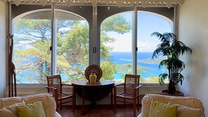 A room with a large window offering a view of the sea and trees. There is a round table with chairs and a potted plant.