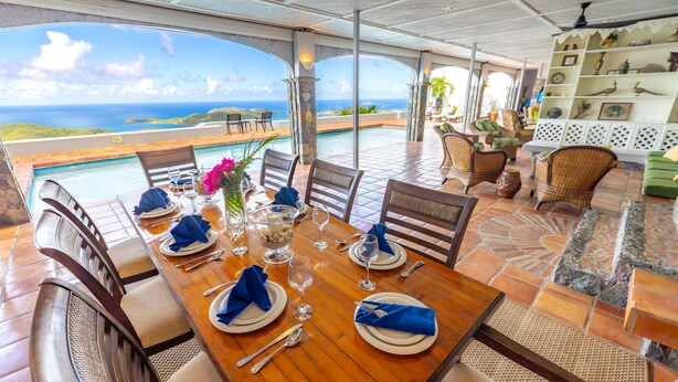 A dining area with a view of the ocean, a pool, and a terrace with wicker furniture and a decorative shelf.