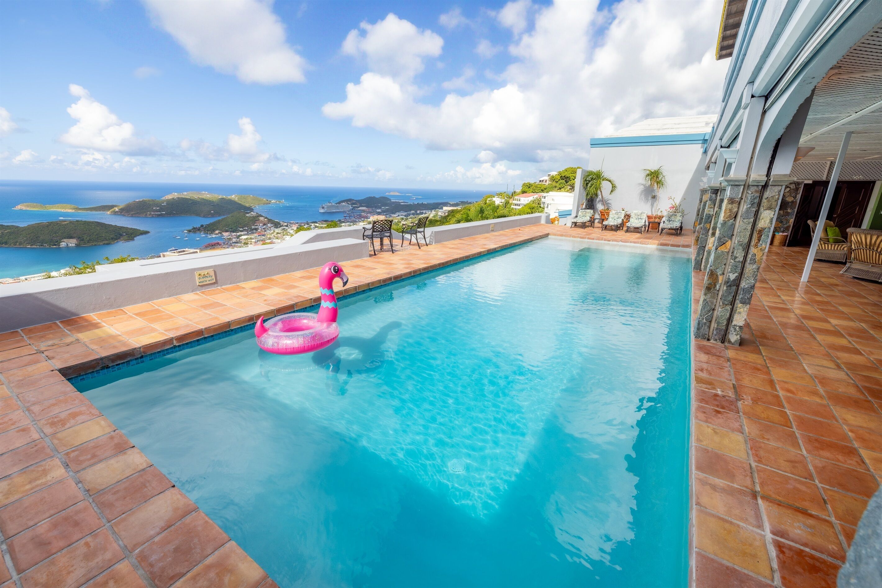 Une piscine avec vue sur l’océan et les collines, un flotteur en forme de flamant rose et du mobilier d’extérieur.