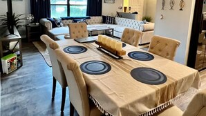 Un coin repas avec une longue table, des chaises et un lustre. On aperçoit un canapé et une petite table avec une lampe au fond.