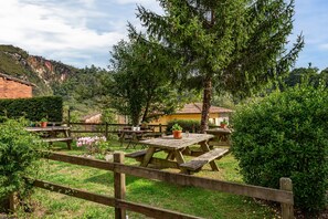 A rustic outdoor dining area with wooden picnic tables, surrounded by greenery and a mountain backdrop.