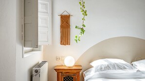 Een slaapkamer met een bed, een nachtkastje met een lamp, een radiator en een macramé wanddecoratie.