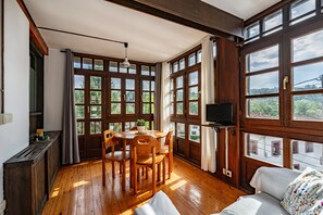 Quarto com piso de madeira, mesa de jantar com cadeiras, sofá e janelas amplas com cortinas.