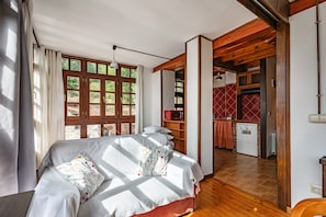 Quarto com uma cama grande, janela com cortinas, área de cozinha com geladeira e teto de madeira.