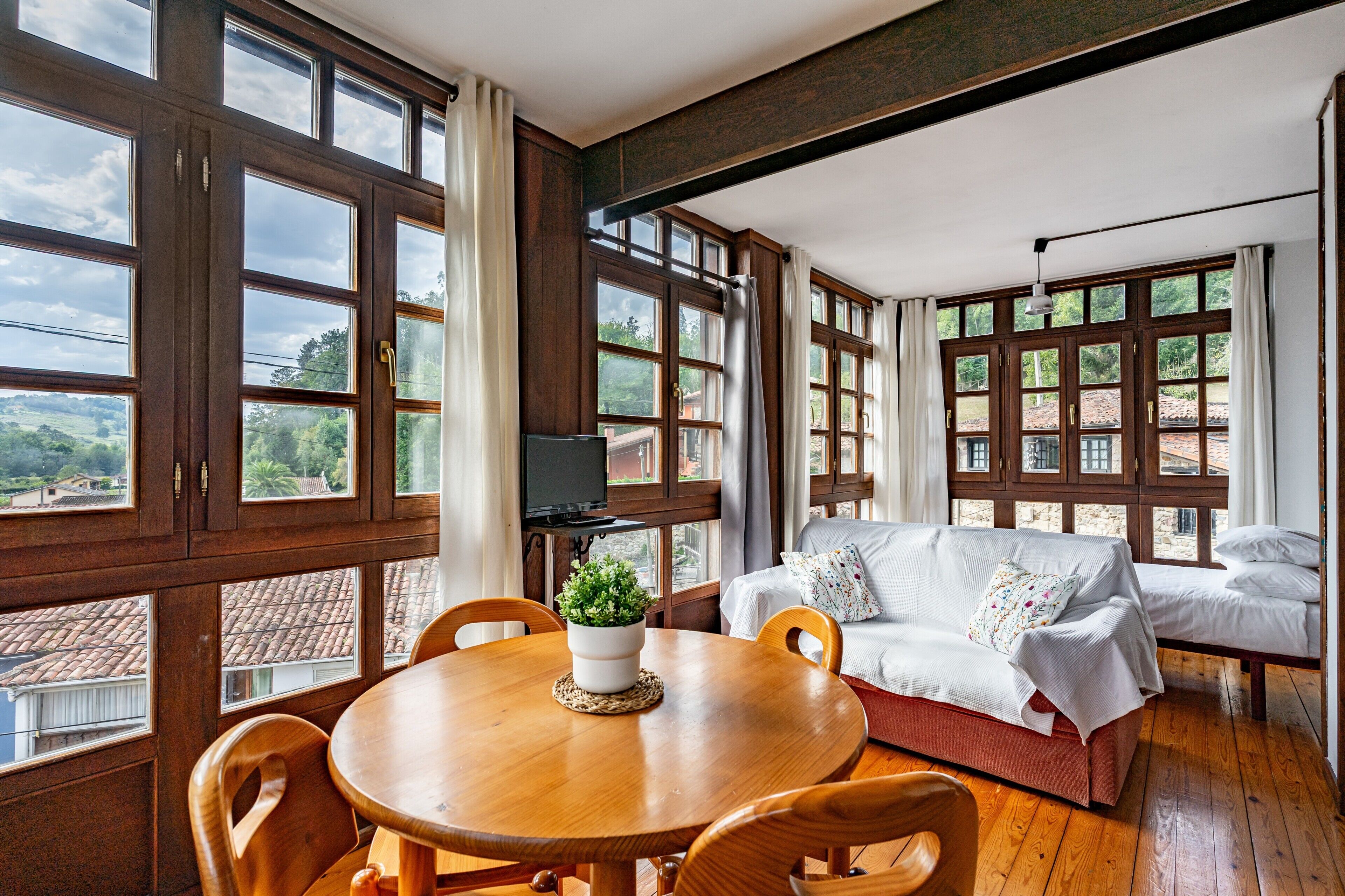 Una habitación con una mesa de comedor de madera, sillas, un sofá y ventanas grandes con cortinas.