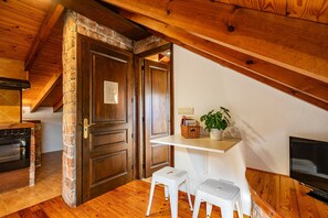 A room with a stone wall, wooden ceiling beams, a brick oven, a white shelf with a plant, and two white stools.