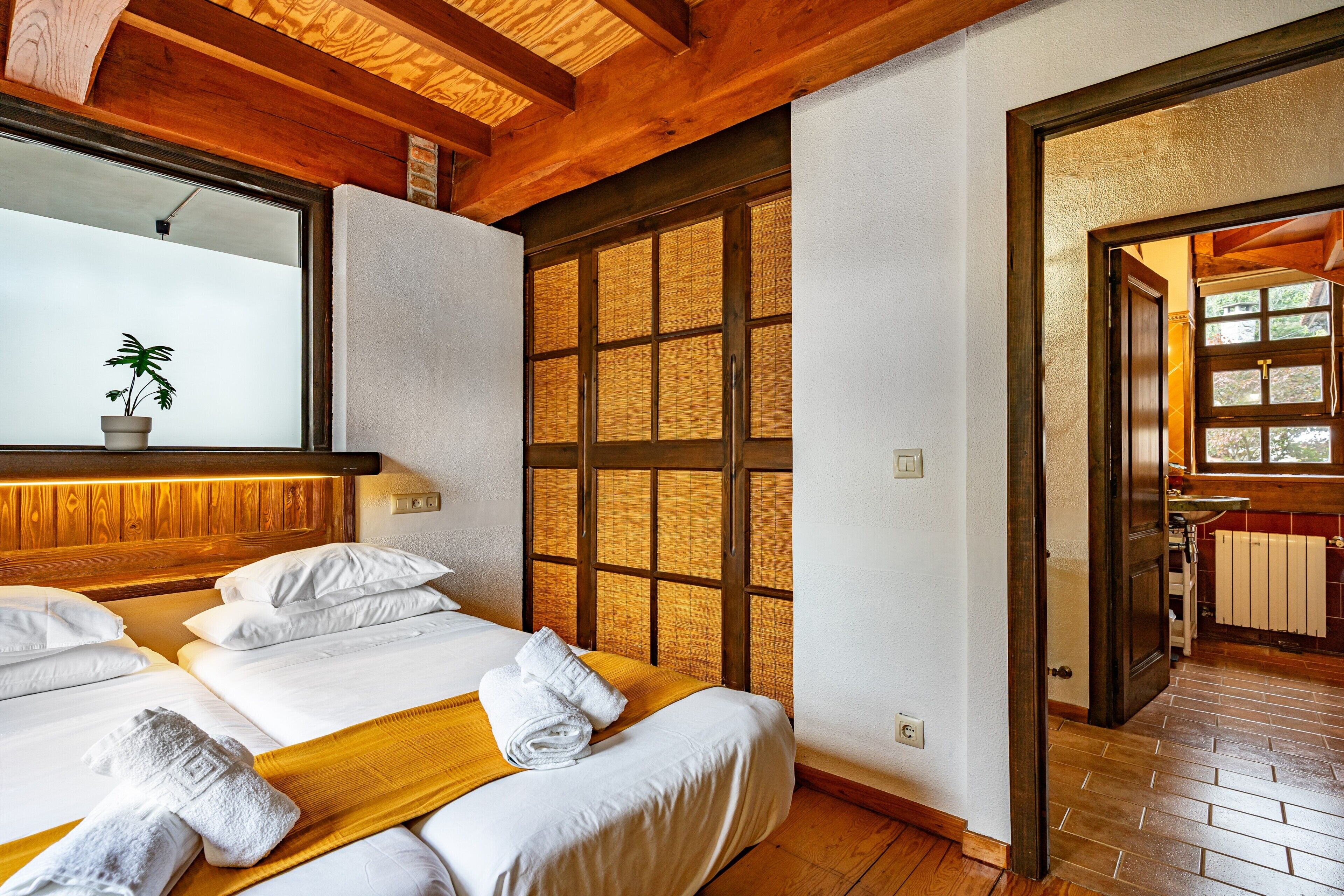 A hotel room with wooden beams, a large window with bamboo shades, a potted plant, and neatly made beds with towels.