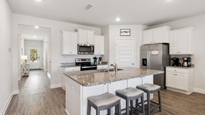 A modern kitchen with white cabinets, a granite countertop, stainless steel appliances, and a central island with bar stools. - Falling Star Retreat is a 4BA/2BA coastal home, just 900 feet to the beach, pet friendly! (Port St. Joe)
