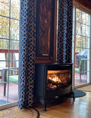 A room with a wood-burning stove, patterned curtains, and large windows with a view of trees.