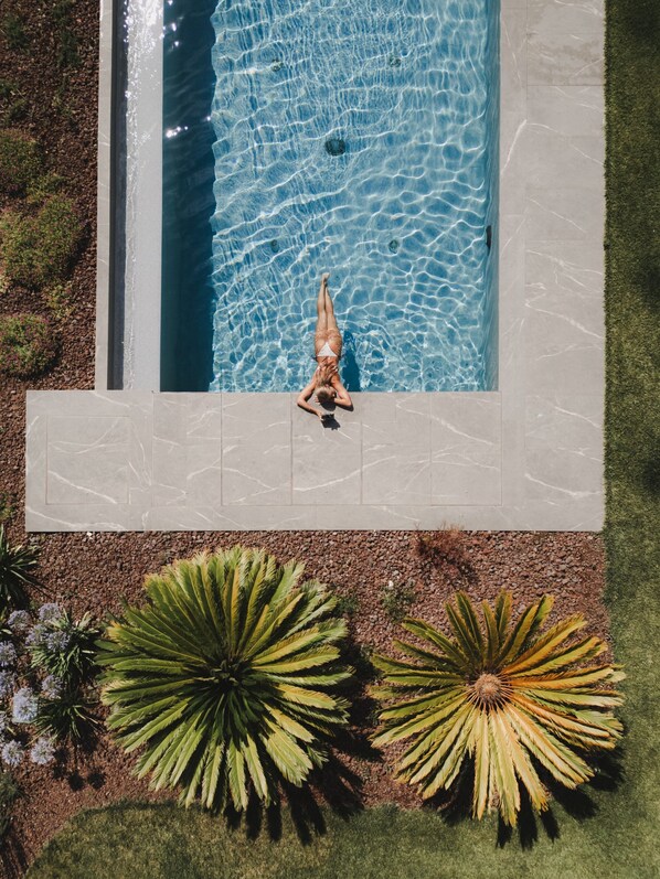 A swimming pool with a person lying on the edge, surrounded by a well-maintained garden with palm-like plants.