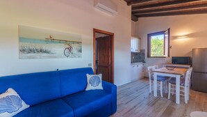 Una habitación con un sofá azul, una mesa de comedor de madera con sillas, una zona de cocina con un refrigerador y un cuadro de una escena de playa.