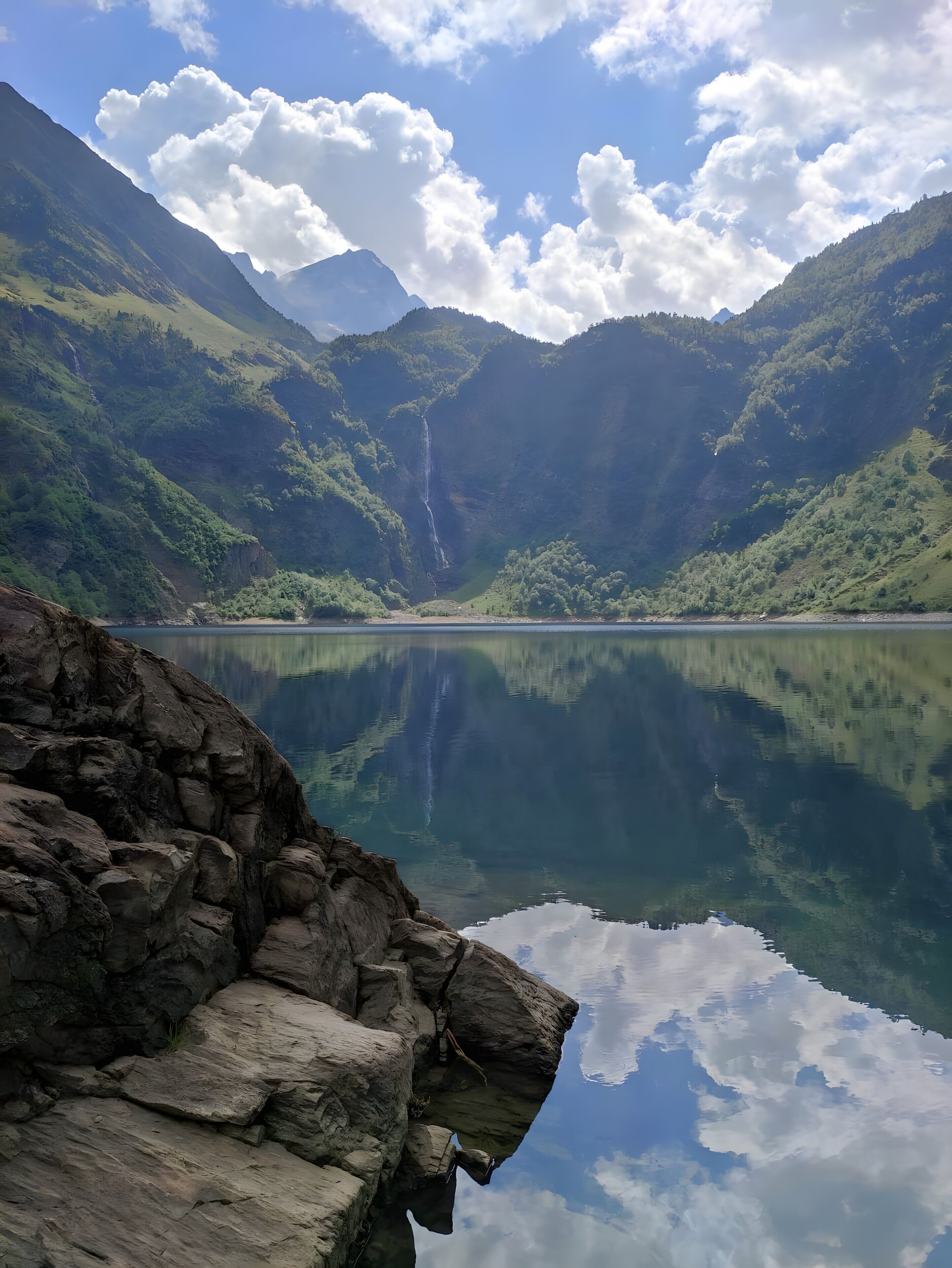 Ein ruhiger See mit einem Wasserfall, umgeben von üppigen grünen Bergen und klarem Himmel.