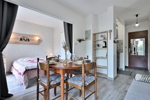 A cozy room with a bed, a dining table with chairs, a shelf with kitchen items, and a wall-mounted light.