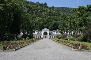 A building with a central entrance, surrounded by a gravel driveway, and lush greenery in the background. - Apartment 'L'Encatador - Bagnères-de-Luchon' with Mountain View (Bagnères-de-Luchon)