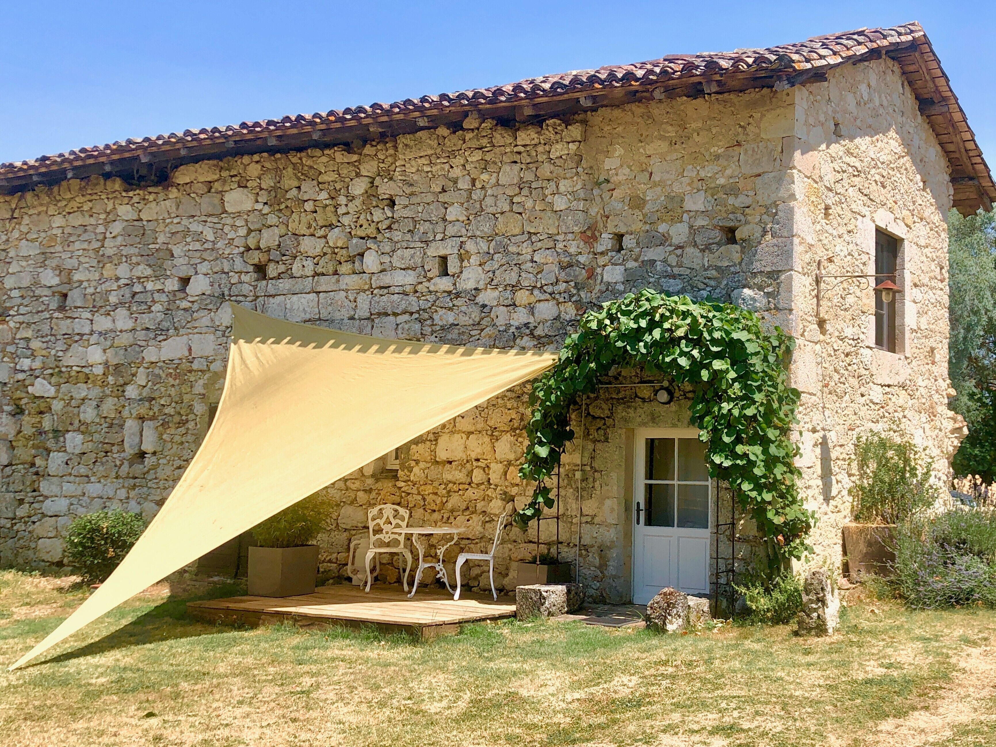 Un bâtiment en pierre avec un toit en tuiles, un espace extérieur abrité avec une table et des chaises, et une porte dotée d’une fenêtre.
