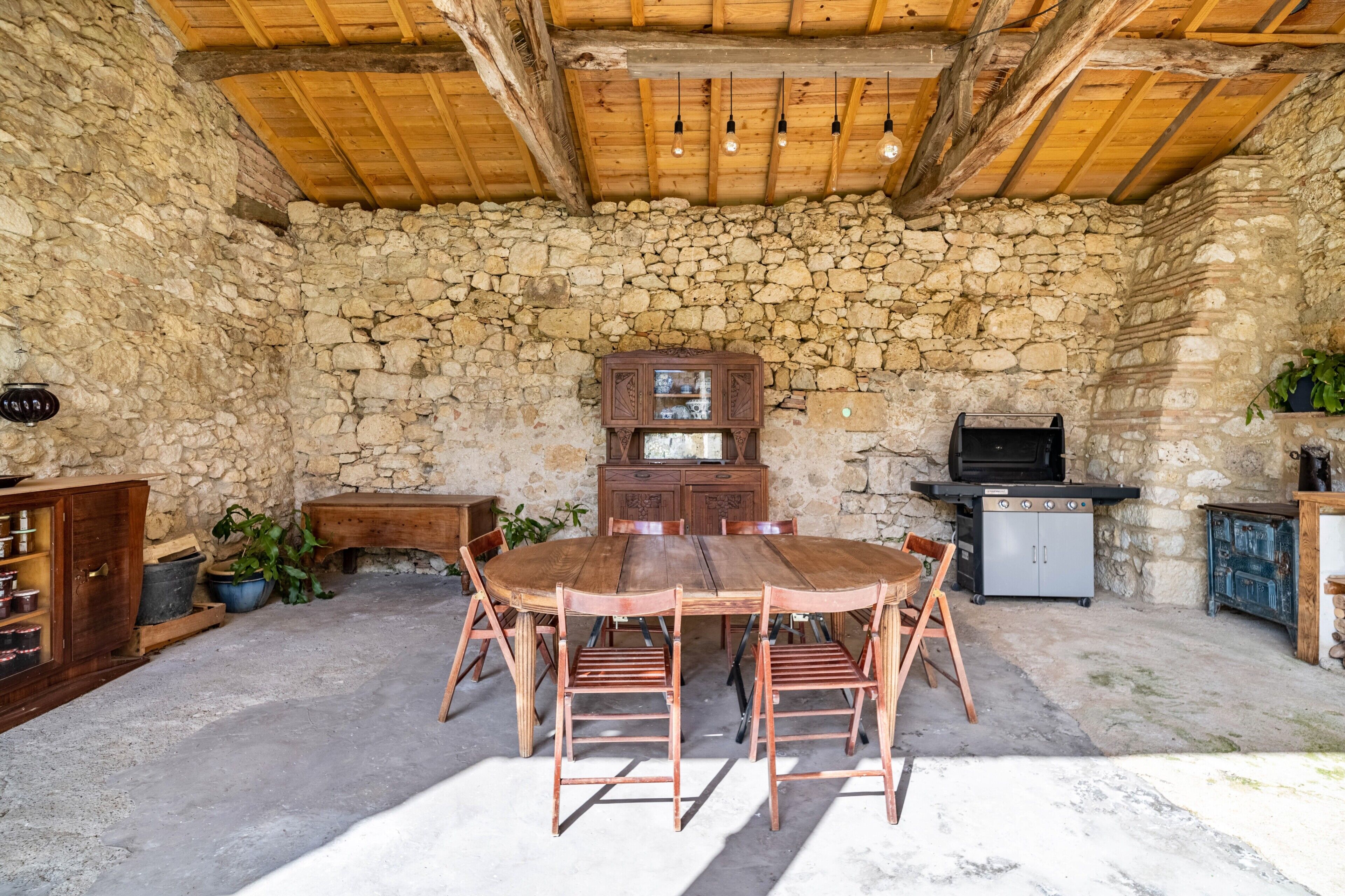 A rustic stone-walled room with a wooden ceiling, a round wooden table with chairs, a cabinet, and a grill.