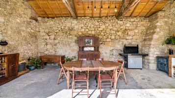 A rustic stone-walled room with a wooden ceiling, a round wooden table with chairs, a cabinet, and a grill.