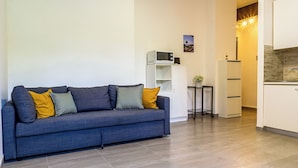 Ein modernes Wohnzimmer mit grauem Sofa, einem kleinen Beistelltisch und einer Küchenzeile mit weißen Schränken und einer Mikrowelle.
