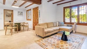 Una sala de estar con un sofá beige, vigas de madera, un tapete estampado y un comedor con sillas de mimbre.