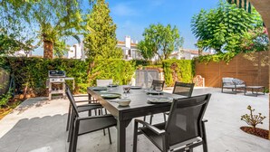 Un patio al aire libre con una mesa de comedor dispuesta para cuatro personas, sillas alrededor y una parrilla al fondo.