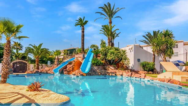 Una piscina con dos toboganes, palmeras y un cielo azul despejado.