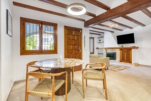 A room with a round wooden table, two wicker chairs, a wooden door, a television, and a fireplace. - Holiday Home 'Ortega' with Shared Pool, Wi-Fi and Air Conditioning (Cala Blanca)