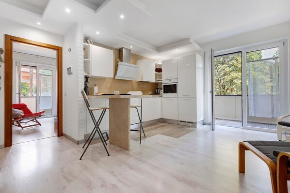 Une cuisine moderne avec des meubles blancs, un îlot central et une porte-fenêtre coulissante donnant sur un balcon.
