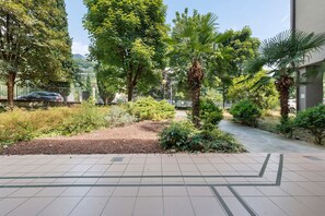 A paved walkway with a palm tree and greenery, a fence, and a building in the background.