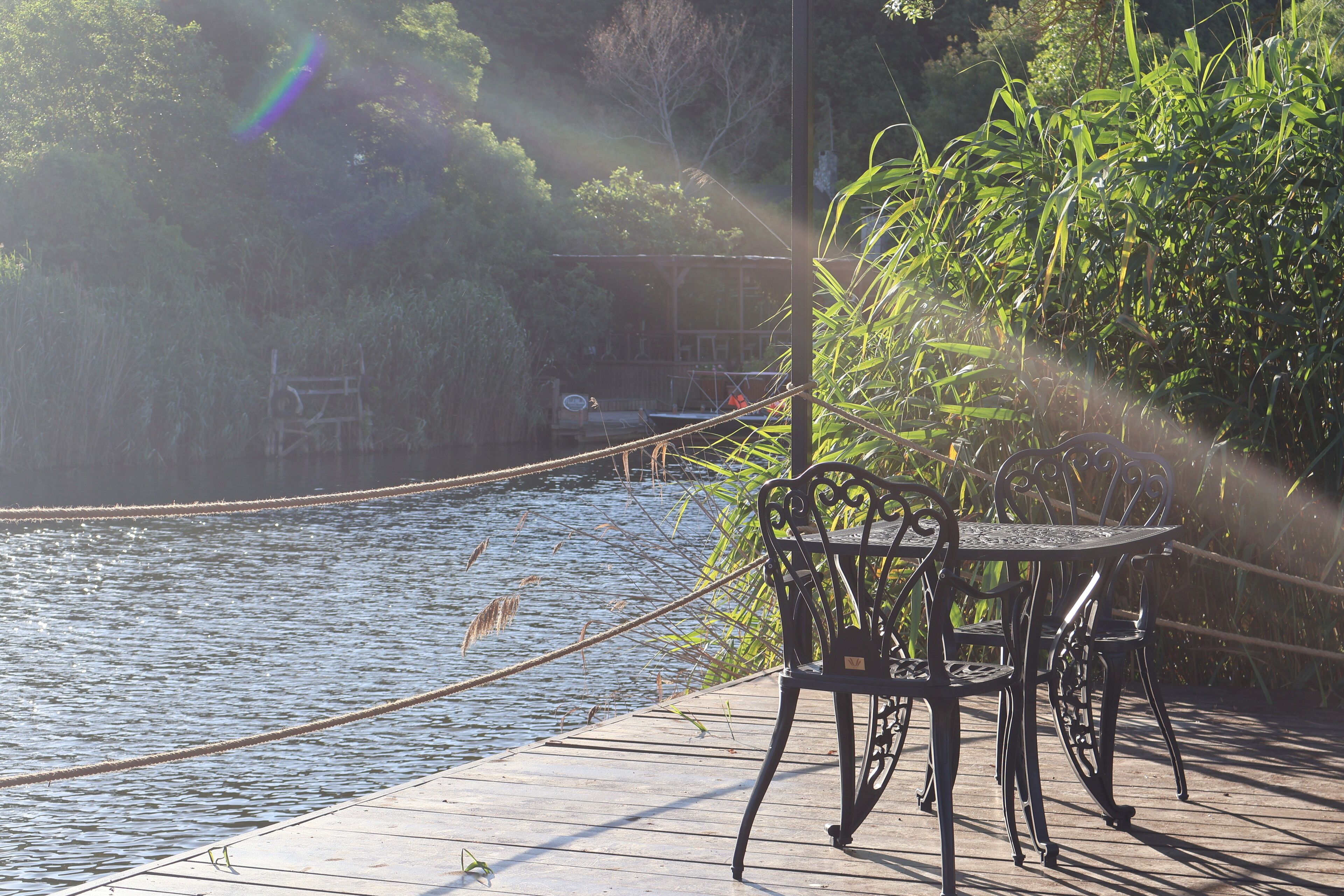 A wooden deck with a metal table and chairs overlooking a body of water, surrounded by greenery.