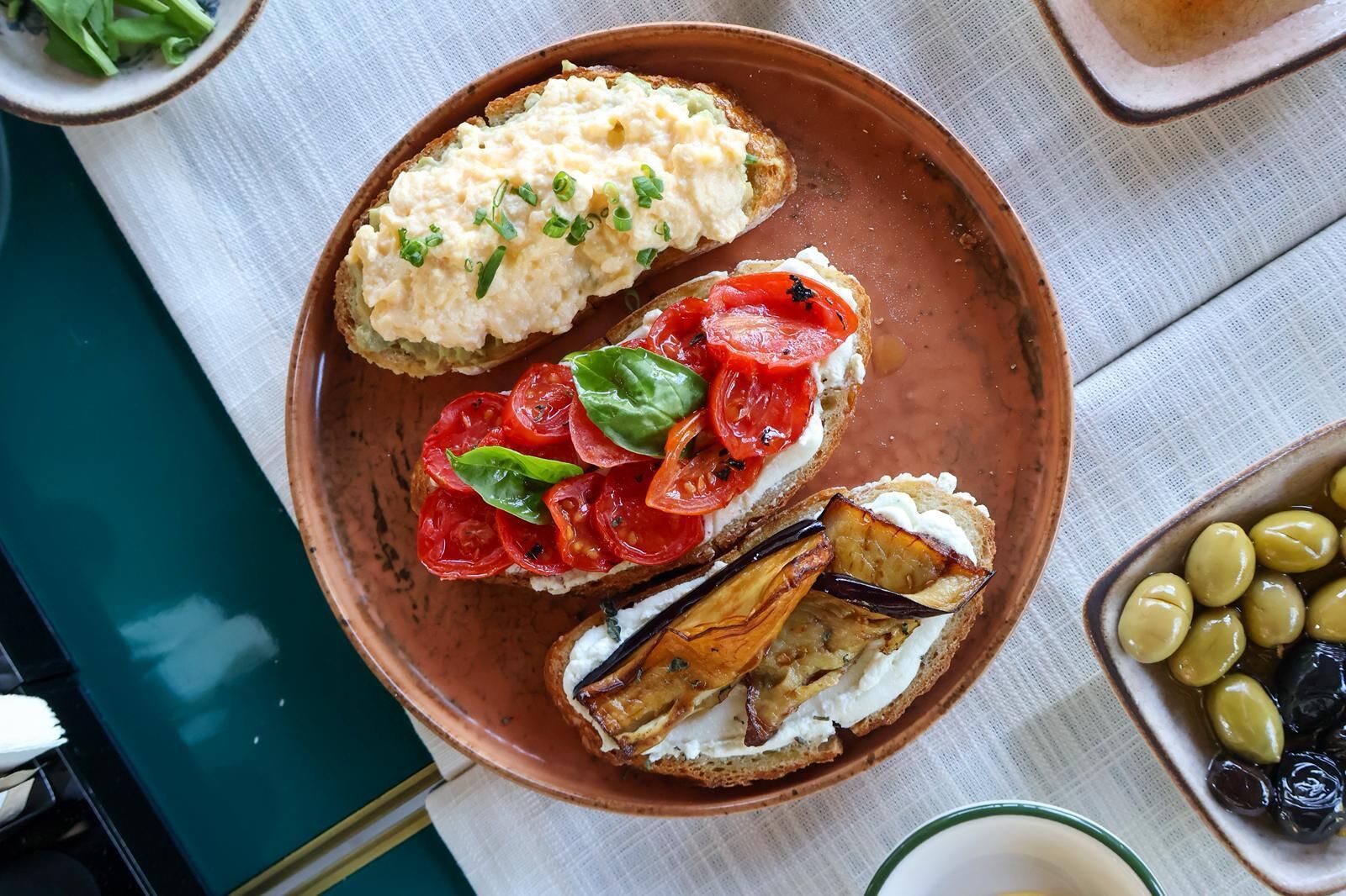 A plate with three open-faced sandwiches, one with a creamy spread, another with tomato slices and basil, and the third with roasted eggplant and a creamy spread.