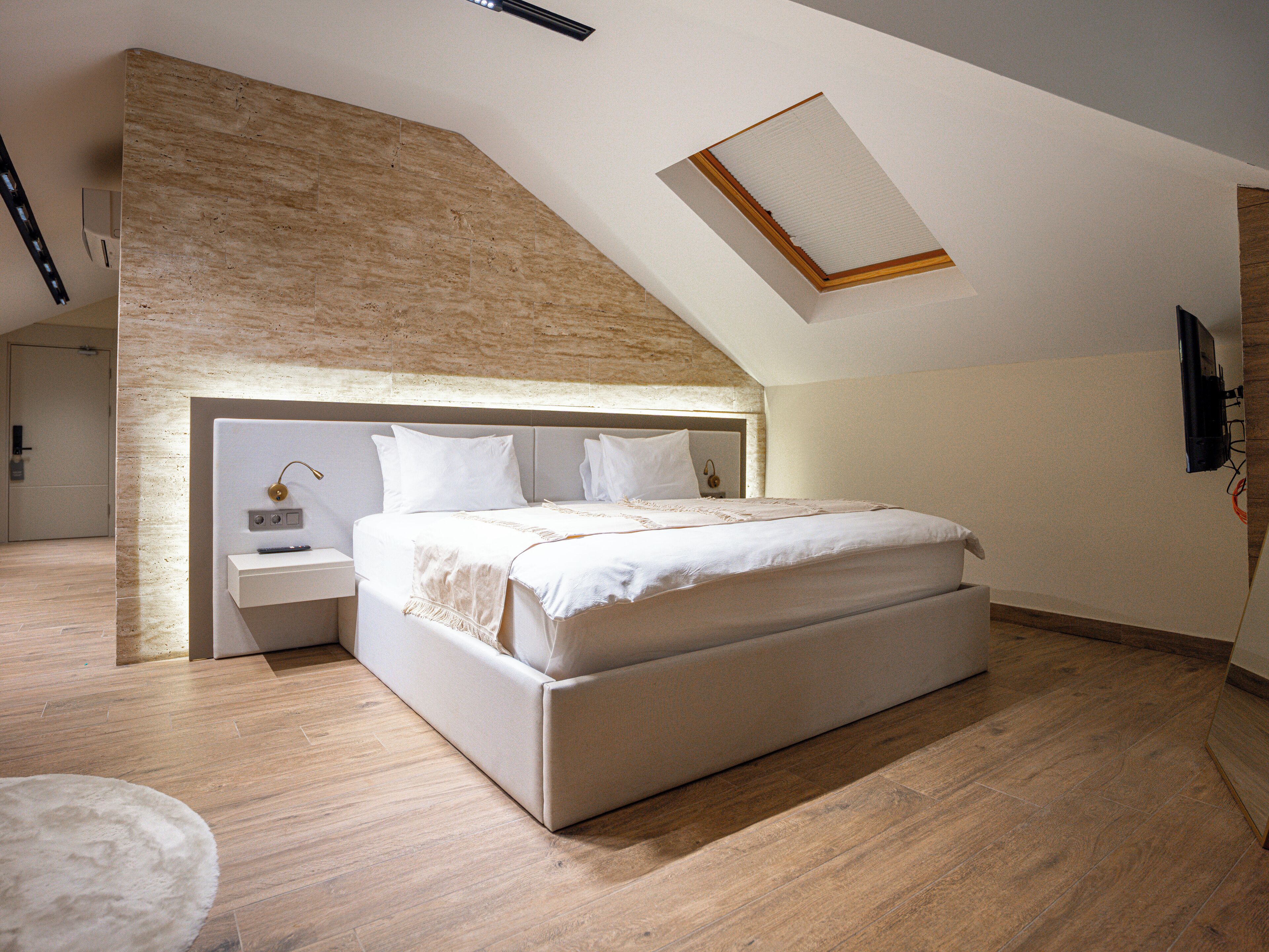 A bedroom with a large bed, a nightstand, a wall-mounted lamp, and a sloped ceiling with a skylight.