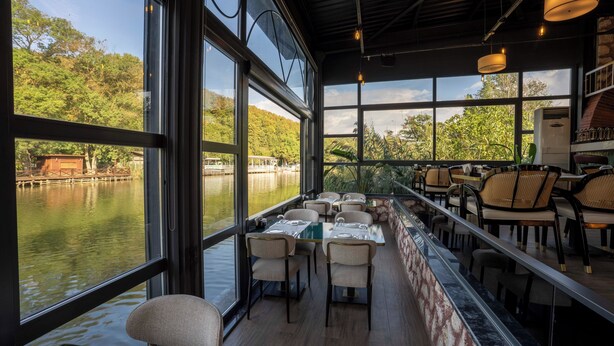 A restaurant with large windows overlooking a lake, wooden deck seating, and a view of boats and greenery. - Verona Hotel (Sile)