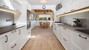 Una cocina con armarios blancos, una mesa de comedor con sillas, una cafetera y un reloj en la pared.