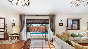 Una habitación con una mesa de comedor, sillas, un sofá con cojines, vista a una piscina y una lámpara de techo.