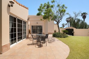 Une terrasse avec une table et des chaises, un espace couvert avec une porte, et une pelouse avec des arbres.