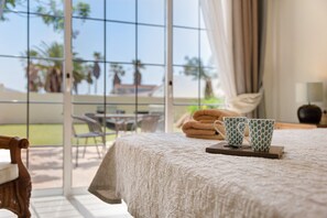 Habitación de hotel con un ventanal grande, cama con cubrecama texturizado, mesita de noche con dos tazas estampadas y vista a palmeras y un edificio.