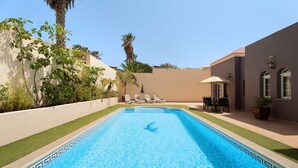 A rectangular swimming pool with a geometric tile border, surrounded by a low wall, a palm tree, and a building with a flat roof.
