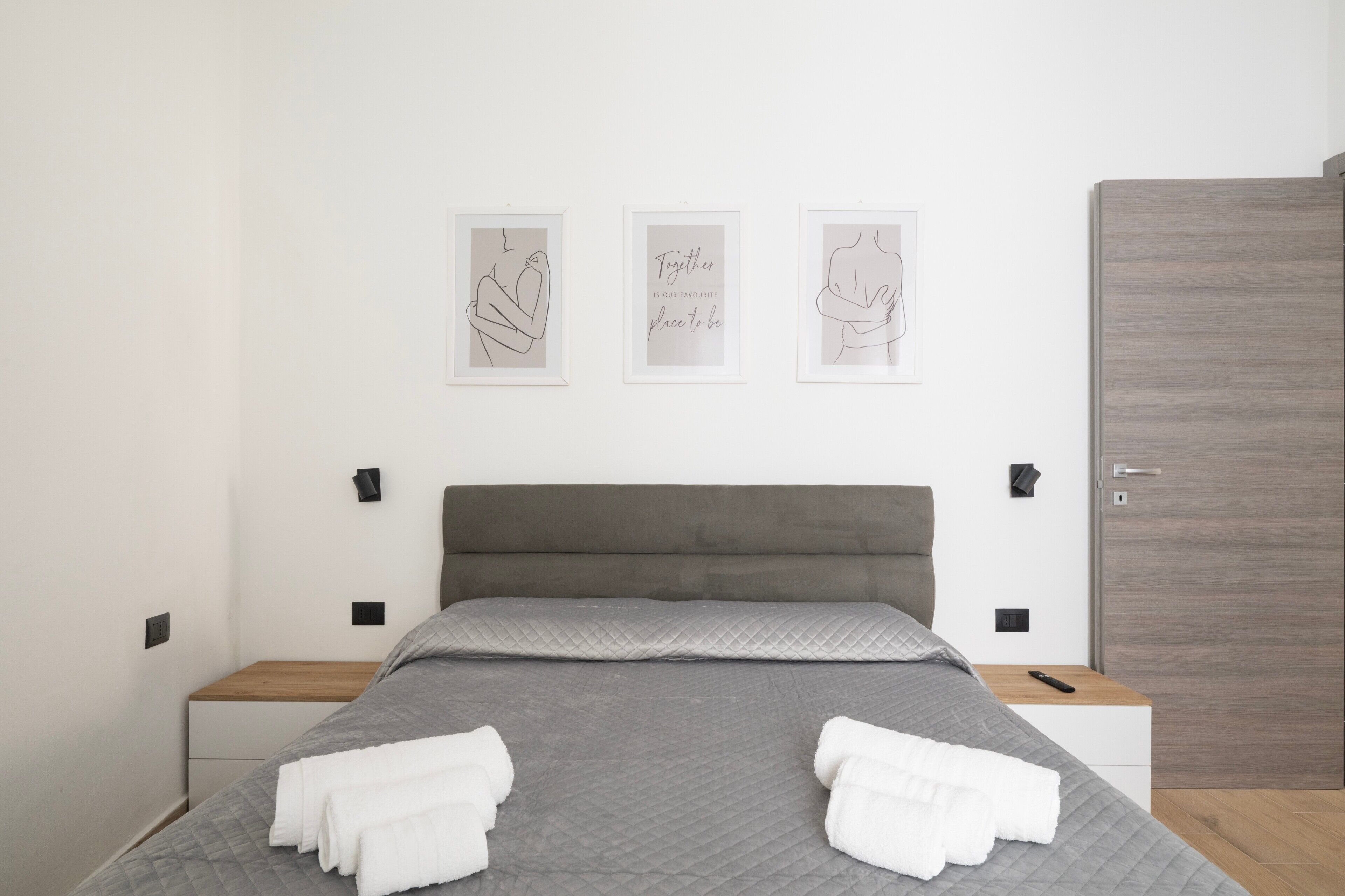 A modern bedroom with a grey quilted bedspread, two white towels, a grey upholstered headboard, and three framed artworks on the wall.