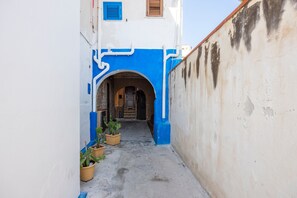 Une ruelle étroite, surmontée d’une arche bleue, bordée de plantes en pot et flanquée d’un immeuble à la façade blanche et bleue.