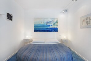 Une chambre à coucher comprenant un lit, deux tables de chevet, un tableau représentant un paysage côtier et un luminaire fixé au mur.