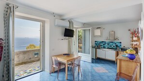Ein Zimmer mit Esstisch, Stühlen, Fernseher, Küchenzeile und Meerblick.
