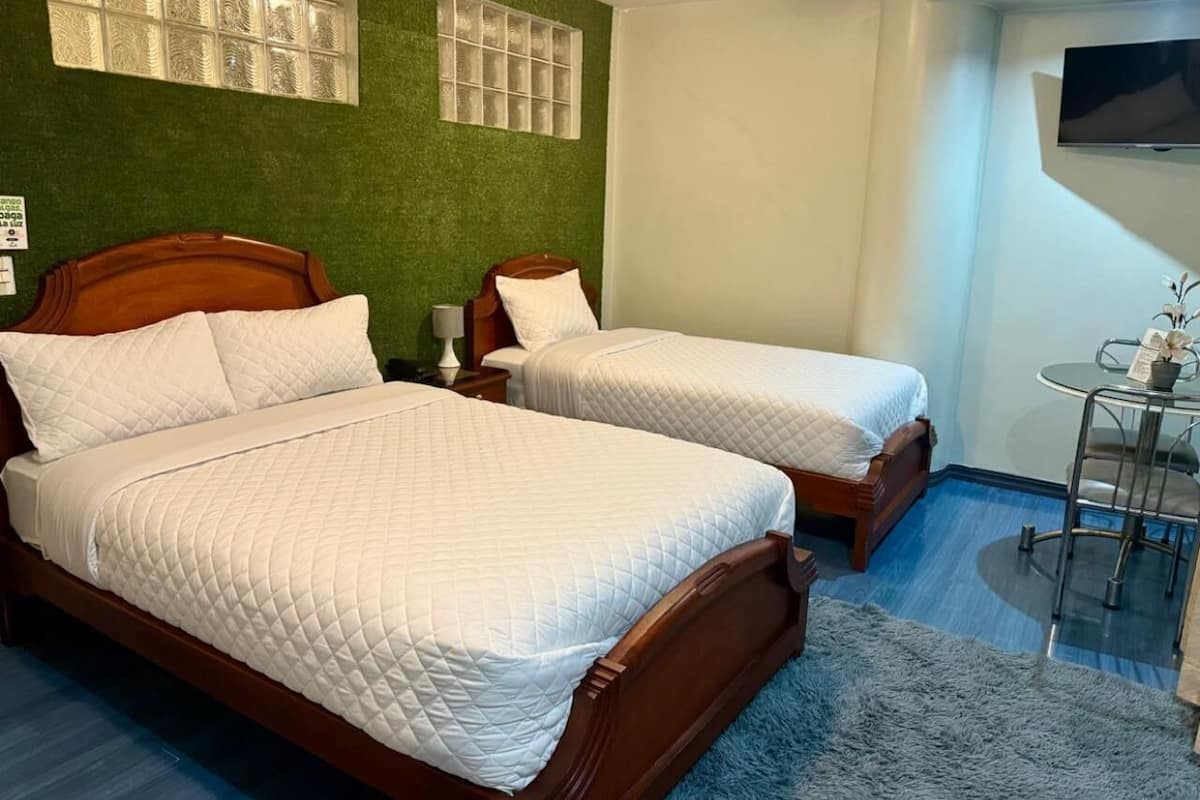 A hotel room with two wooden beds, a green accent wall, a TV mounted on the wall, and a small round table with a chair.
