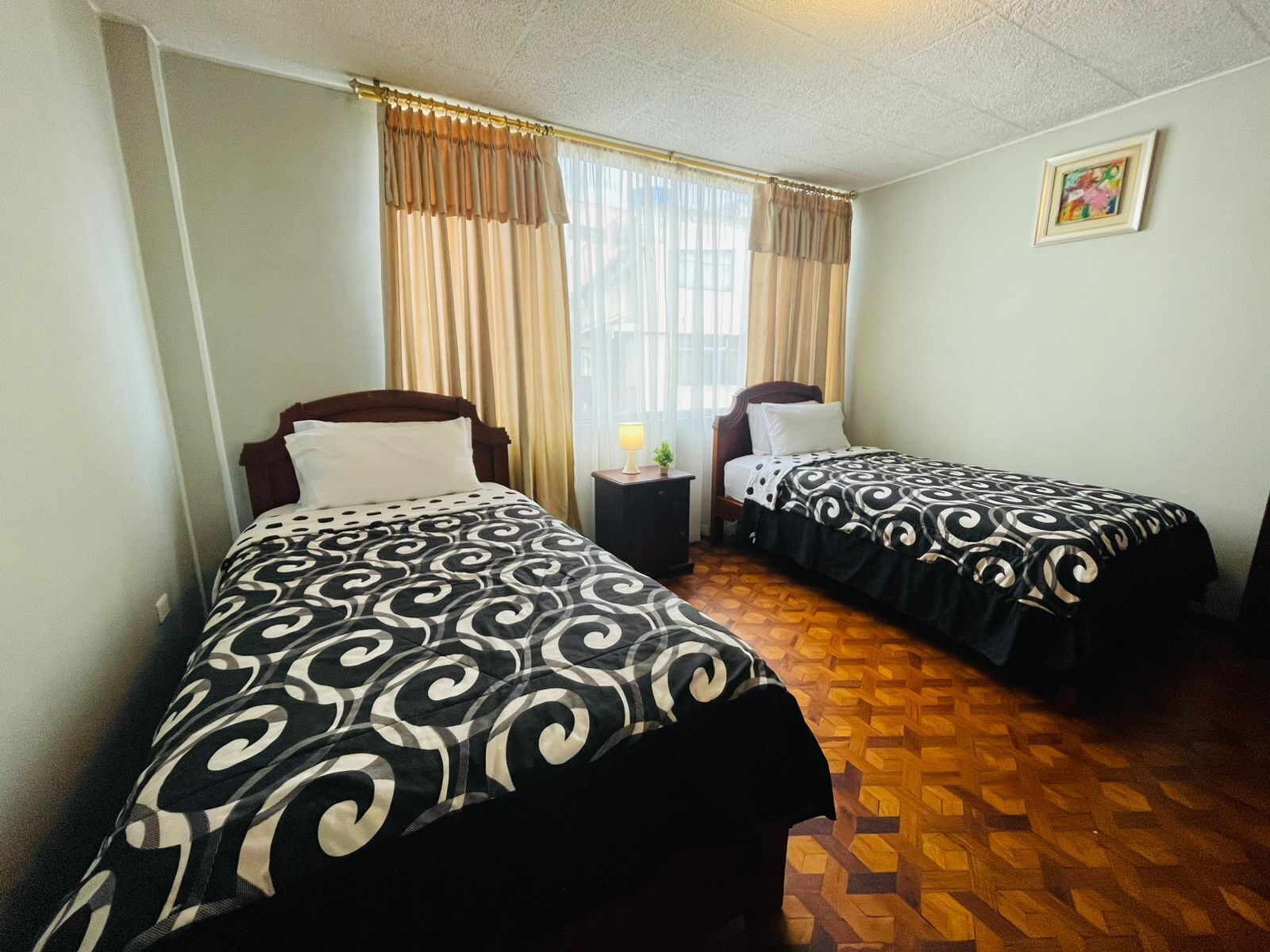 Habitación de hotel con dos camas individuales, una mesita de noche con lámpara, una ventana con cortinas y un cuadro en la pared.