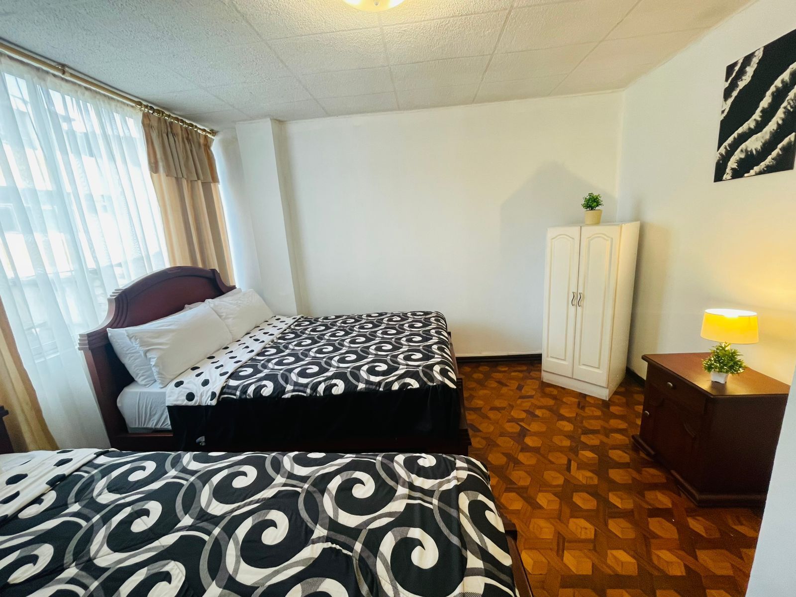 Una habitación de hotel con cama, armario, mesita de noche con lámpara y ventana con cortinas.