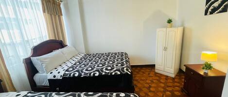 Una habitación de hotel con cama, armario, mesita de noche con lámpara y ventana con cortinas.
