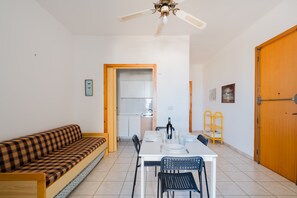 Ein Zimmer mit einer Couch, einem Esstisch, Stühlen, einem Deckenventilator, einer Holztür und einer Küchenzeile.