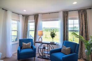 A room with two blue armchairs, a small table with a lamp, and a view of greenery outside.