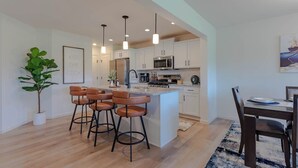 Una cocina moderna con una isla central, taburetes de bar, iluminación colgante y un comedor con mesa y sillas.