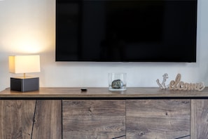 A flat-screen TV mounted on a wall, a lamp on a wooden console, a glass vase with a decorative item, and a 'Welcome' sign.